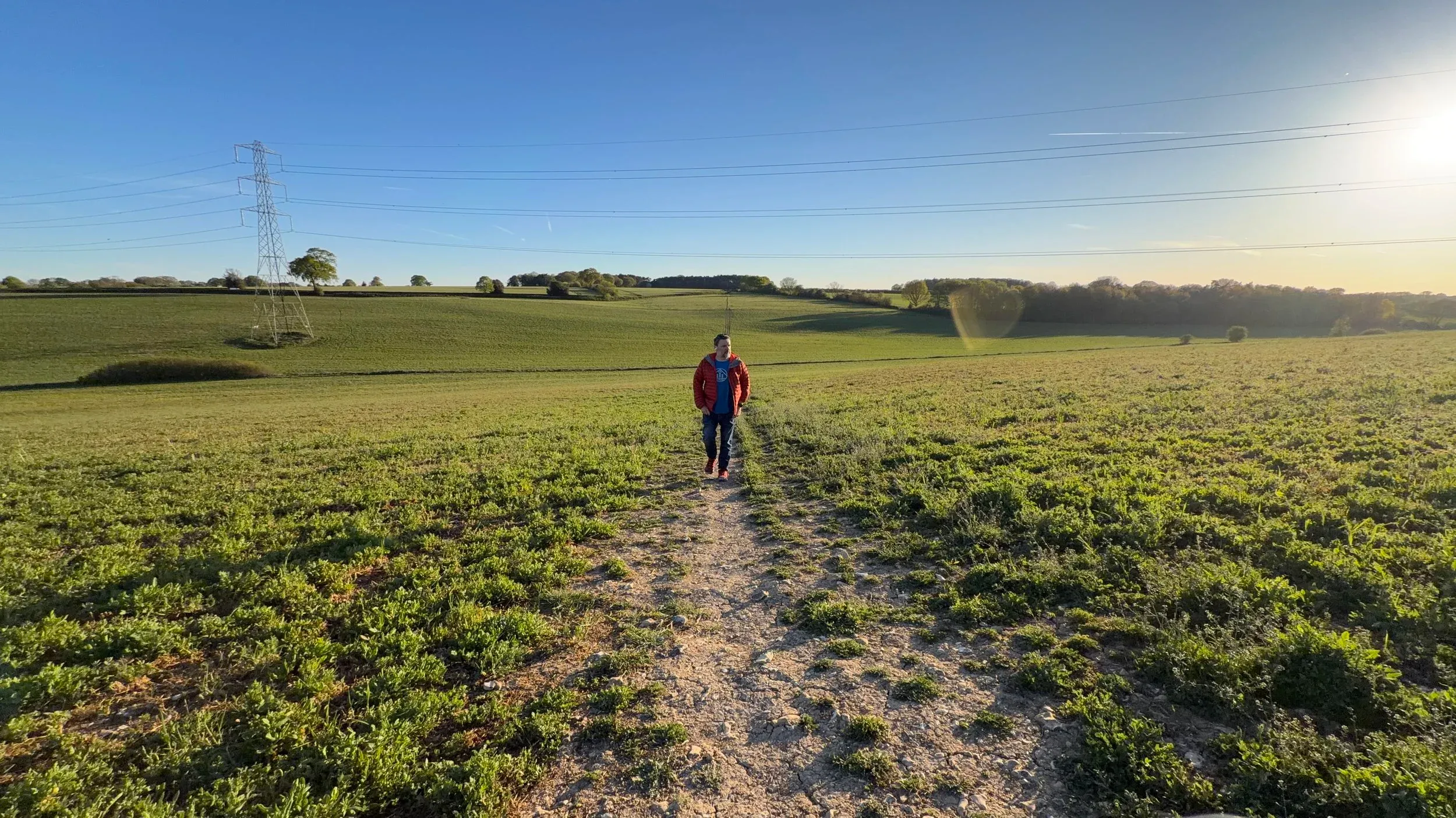 A person walking a dirt path towards camera across an open green field, low sun flaring behind
